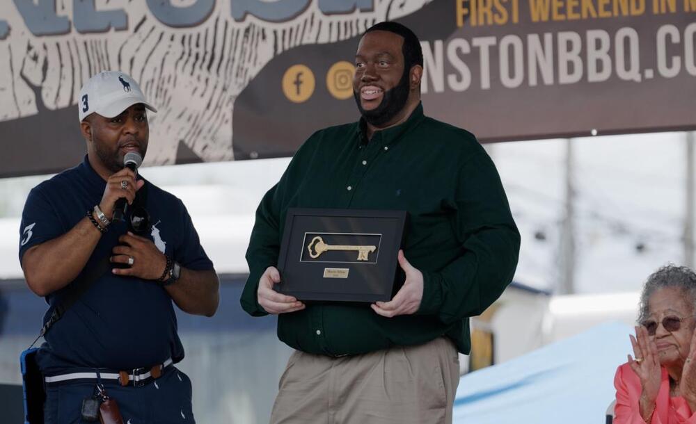 Man with a microphone speaking while a man holds a key to the city. A woman applauds behind them. 