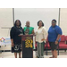 A group of women stand around the award recipient, behind the podium with the 2024 decoration on it. 